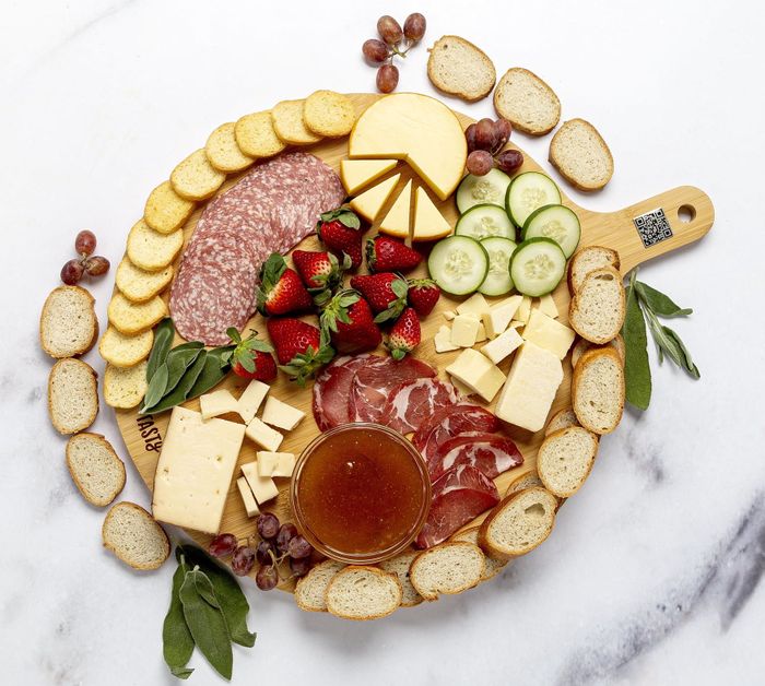 A round bamboo charcuterie board with a handle, filled with a variety of cheeses, salami, strawberries, cucumber slices, prosciutto, grapes, and a honey dip, alongside fresh baguette slices.