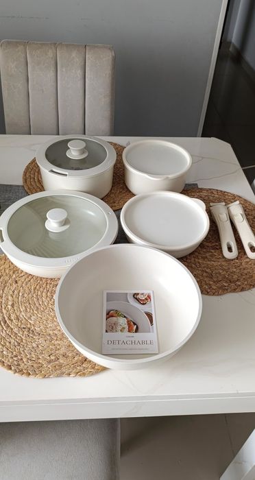 A beautifully arranged table setting featuring cookware: two lidded pans, two bowls on placemats, three detachable utensil handles, and a cookbook titled 