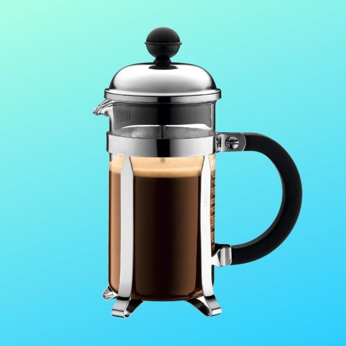 A French press sitting next to a cup of coffee, set against a smooth blue gradient. It features a minimalist, modern design with a sleek stainless steel body and a black handle.