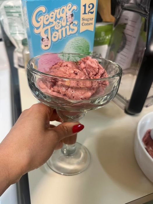 A hand with red nail polish holds a dessert dish filled with ice cream in front of a George & Tom's sugar cone box