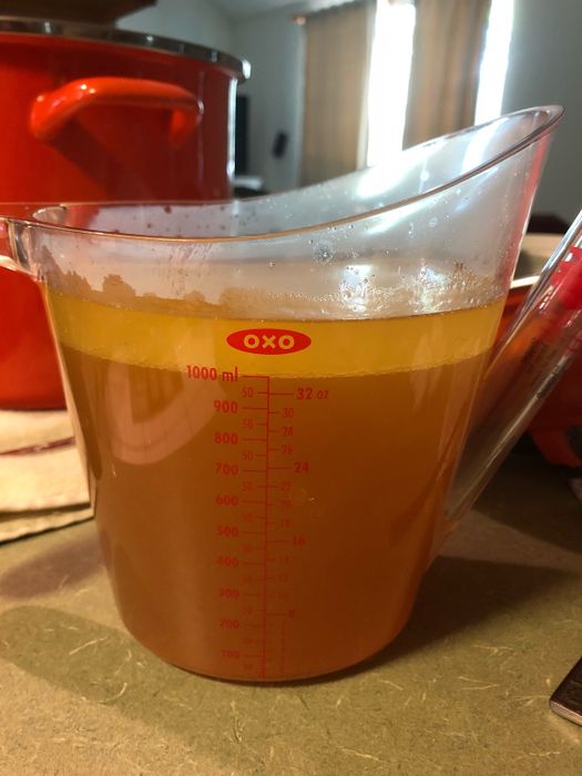 A clear measuring cup filled with broth on a kitchen counter, with a pot and curtains in the background