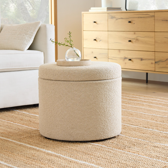 A charming storage ottoman upholstered in soft, light-colored tweed fabric, topped with a small vase holding a delicate plant.
