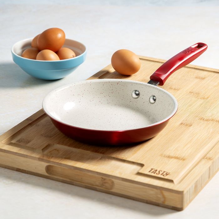 A small nonstick frying pan with a red handle rests on a wooden cutting board beside a bowl of eggs.