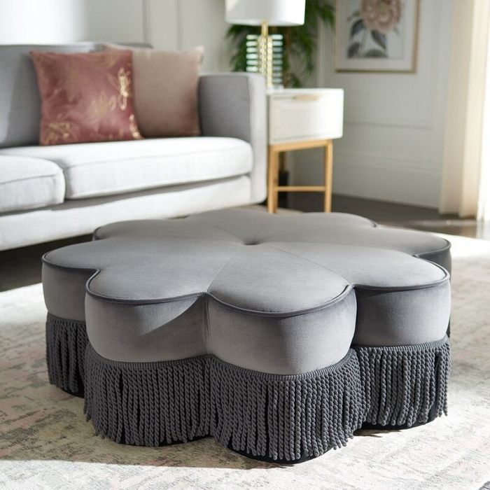 A graceful ottoman with a petal-like shape and fringe accents, placed in a chic living room featuring a sofa and decorative pillows