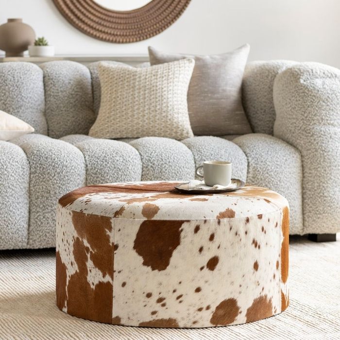 A cozy living room featuring a textured gray sofa, beige throw pillows, and a round cowhide ottoman, with a tray holding a cup resting on top.