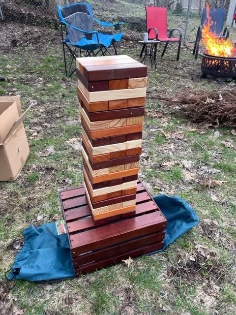 A large outdoor Jenga set neatly stacked on a wooden crate, surrounded by lawn chairs and a fire pit, set in a grassy, leaf-covered backyard.
