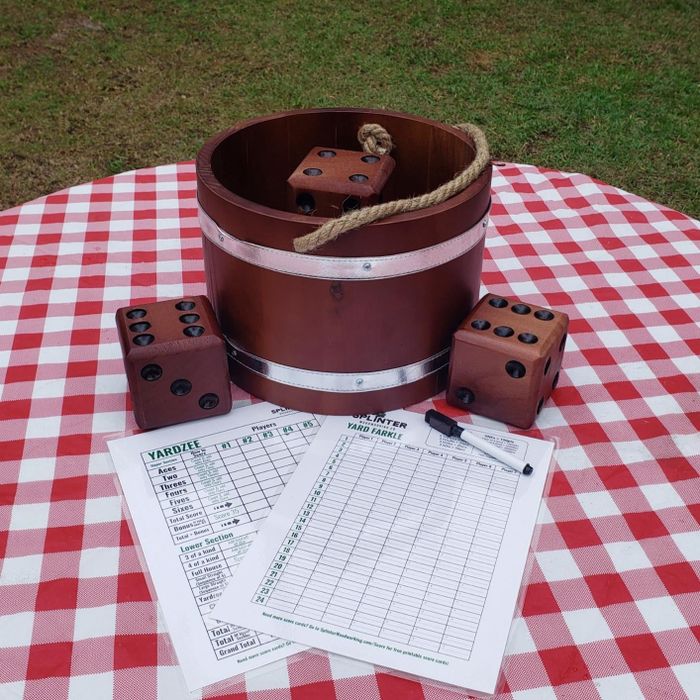 A Yardzee game set placed on a checkered tablecloth, featuring large wooden dice, a bucket, score sheets, and a dry-erase marker