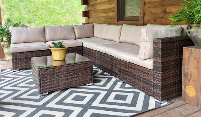 A stylish outdoor patio set with a sectional sofa featuring beige cushions and a glass-topped coffee table, all set on a geometric-patterned rug, placed on a wooden deck