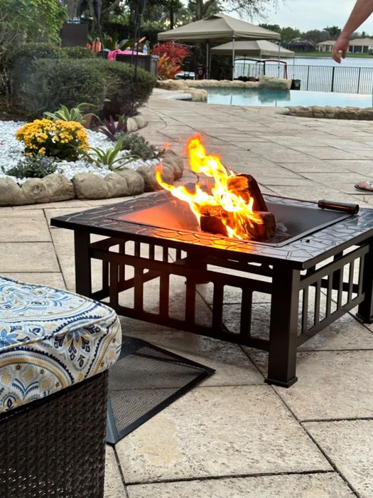An outdoor fire pit on a patio next to a pool, with patterned seating in the foreground. No people visible.