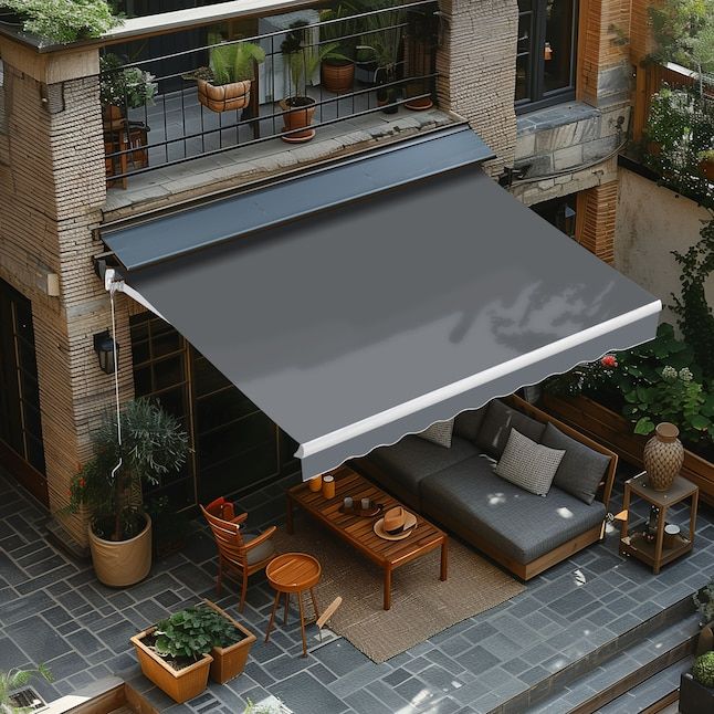 A retractable awning fully extended over a cozy outdoor patio, featuring a gray sofa, wooden table, plants, and outdoor decor. Two stories of windows are visible in the background.