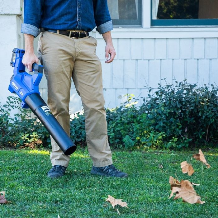 A person operating a leaf blower, clearing fallen leaves from a lawn, with a window and shrubs in the background. The person is dressed in khaki pants, a dark belt, dark shoes, and a rolled-up long-sleeve shirt.