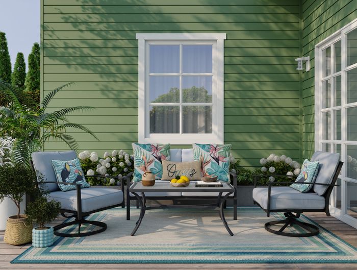 An outdoor patio with green siding, featuring chairs adorned with tropical-themed cushions, a coffee table decorated with lemons and a vase, and several potted plants for extra charm.