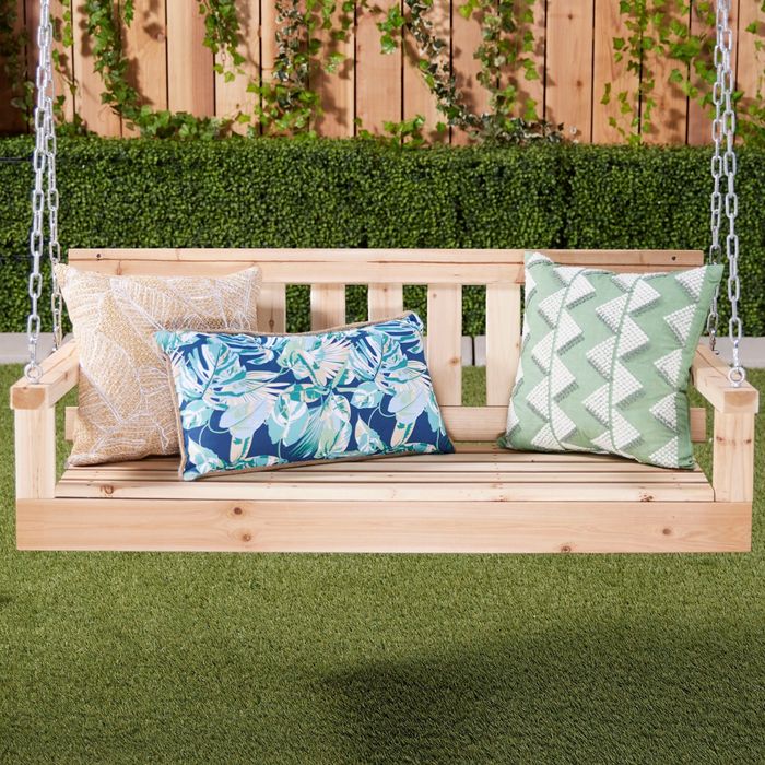 A wooden swing bench set in a garden, with three decorative cushions: one in beige, one featuring a tropical blue design, and one with a green zigzag pattern.