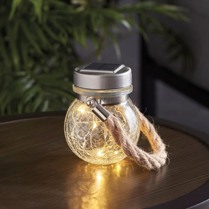 A charming round glass jar with a metal lid and a rope handle, filled with small string lights, rests on a wooden surface.