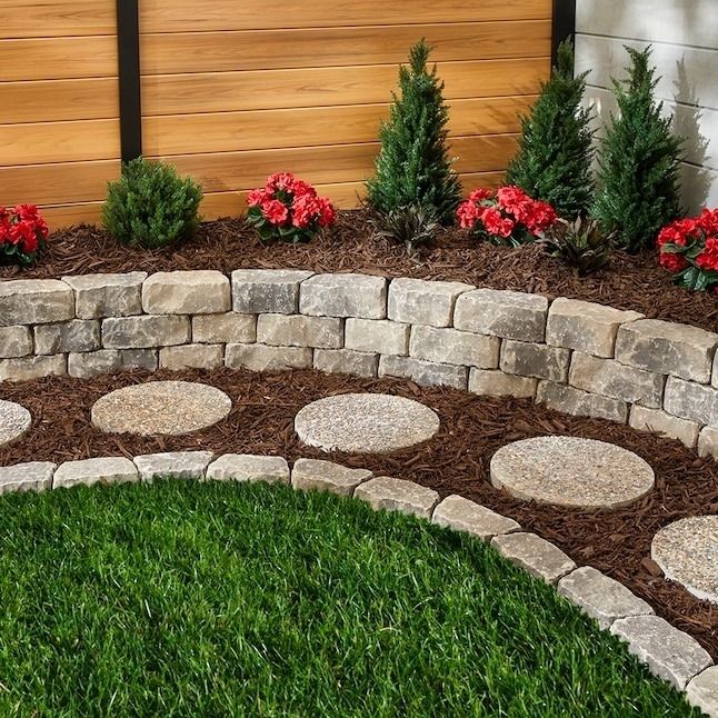 A stone pathway features circular stepping stones, bordered by a curved stone wall with a mulched planting area filled with small shrubs and bright red flowers.