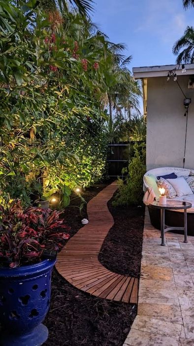 A serene backyard scene with a curved wooden pathway, softly illuminated by garden lights, surrounded by tropical plants, with a cushioned outdoor chair and small table with a candle