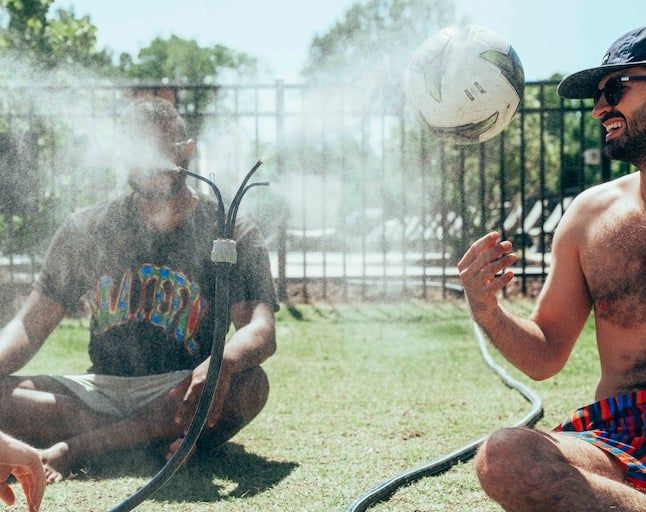 Two unidentified men sitting on the grass next to a sprinkler. One is shirtless, while the other wears a bright T-shirt. Both casually interact with a soccer ball between them.