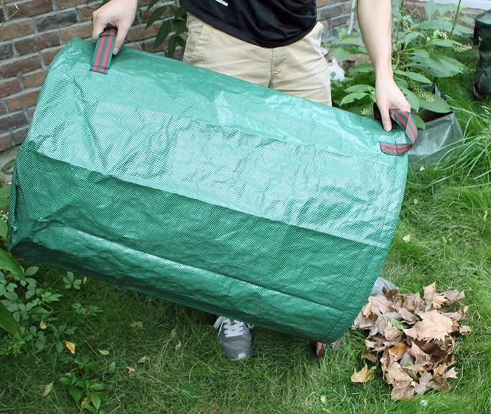 A pair of hands gripping a green cylindrical bag, using it to scoop up dead leaves.