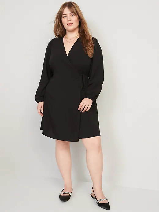 Model wearing a black knee-length wrap dress with long sleeves, a tie waist, and a V-neckline, paired with black pointed-toe flats against a gray background.