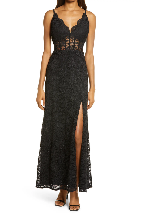 Model showcasing a corset-style black lace maxi dress with a sheer bodice and a leg slit, paired with black open-toed heels.