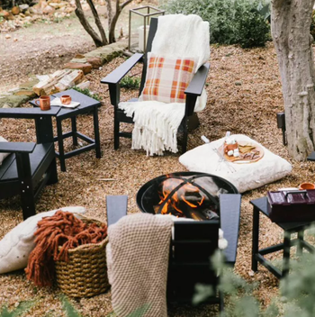A cozy outdoor seating arrangement featuring a fire pit surrounded by chairs, soft blankets, and a plaid pillow, creating a welcoming space for social gatherings.
