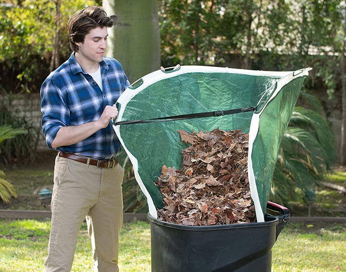 A person uses a tarp to guide leaves into a trash can.