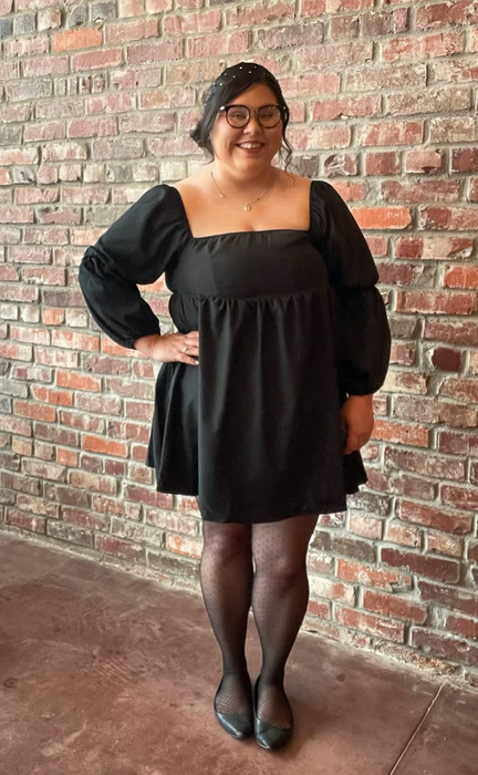 Reviewer in a black puff-sleeved formal dress, paired with black tights and flats, standing in front of a brick wall.