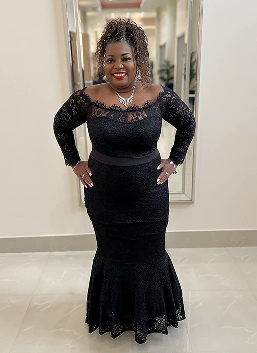 A reviewer is wearing an off-the-shoulder lace dress with a black ribbon tie around the waist, standing in front of a mirror with a white wall behind them.