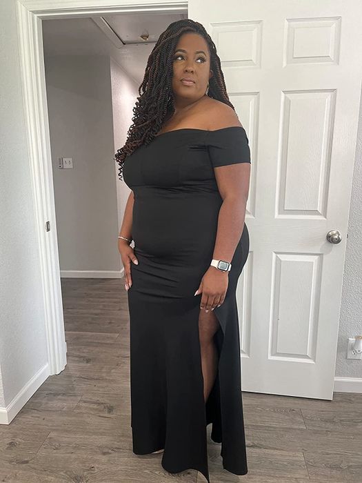 A reviewer in a black off-the-shoulder mermaid dress with a side slit, posing in front of a white door and matching walls.