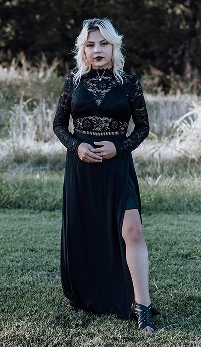 Reviewer wearing a black maxi dress featuring a sheer lace top, long sleeves, and a leg slit, paired with black heels, standing in grass.