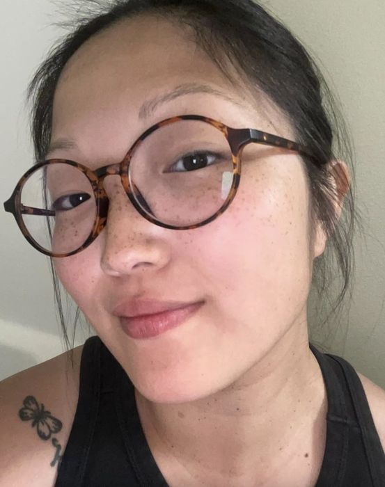 A woman with dark hair tied back, wearing round glasses and a sleeveless top, displays a butterfly tattoo on her shoulder as she gazes at the camera
