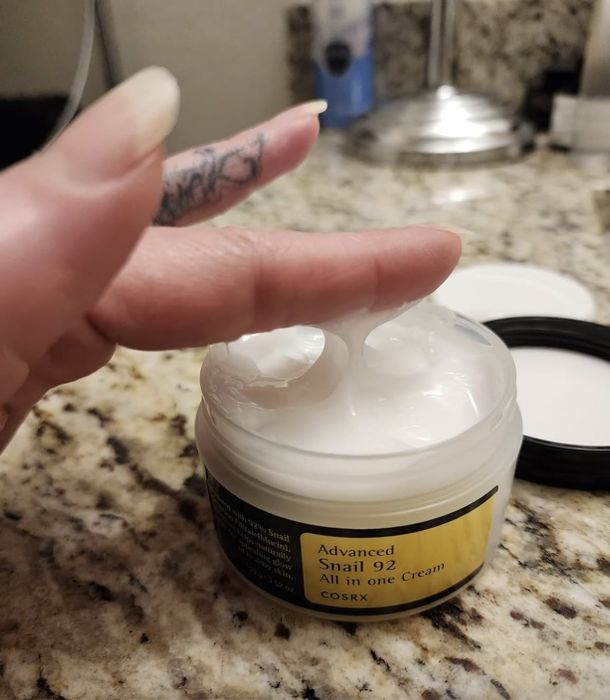 A tattooed hand dipping a finger into an open jar of COSRX Advanced Snail 92 All in One Cream placed on a granite surface