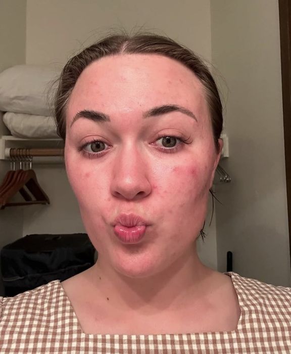A woman wearing a checkered top, her hair tied back, poses with a playful expression in a room filled with a clothing rack and towels in the background