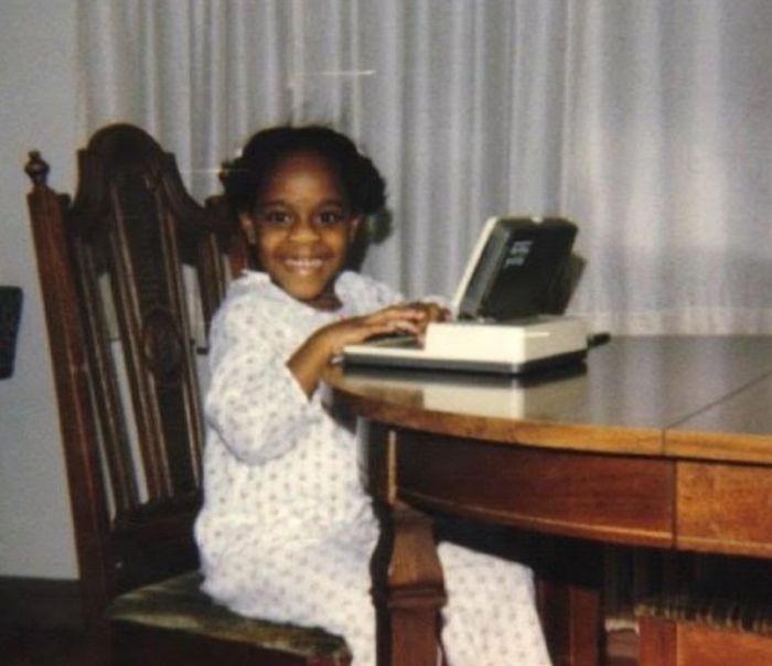 A child smiles at a table with an early laptop-style device, wearing patterned pajamas.