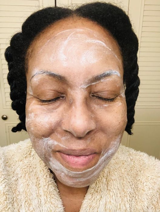 A person with closed eyes, wearing a white facial mask and a plush robe, smiles softly. Their hair is styled in twists, and no names are mentioned