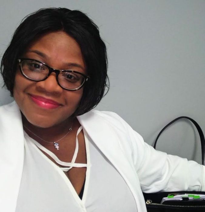 A person smiles, wearing glasses and a white blazer, seated with a black bag beside them.