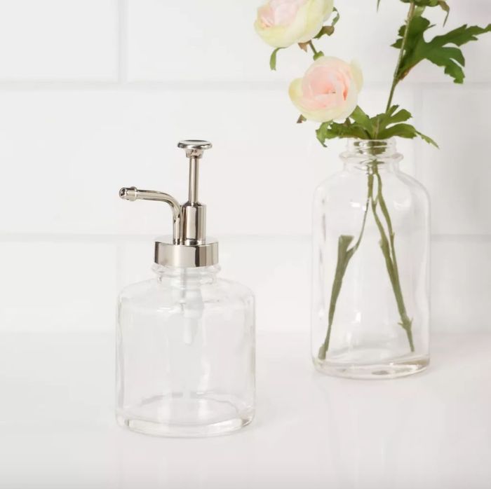 An empty glass soap pump sitting on a counter beside a clear vase filled with flowers.