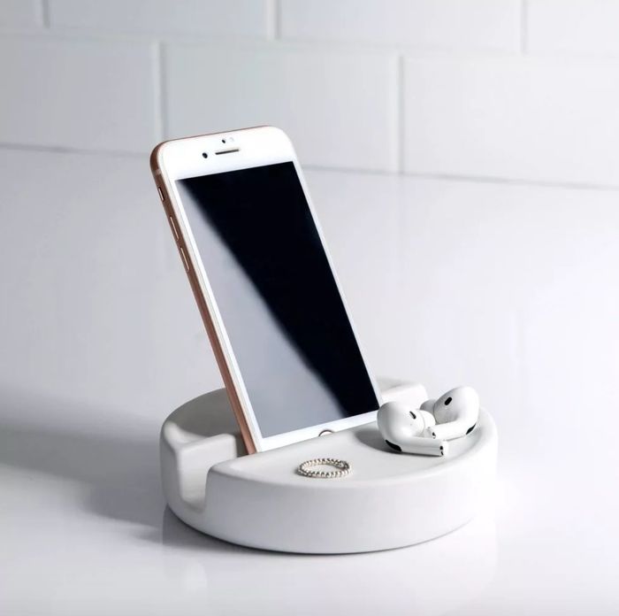 A white, round cellphone holder with a white phone placed on it, accompanied by a ring and AirPods resting on the holder.