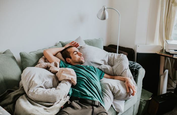 A person lying on a couch, eyes closed, relaxed, surrounded by cushions and blankets in a cozy living room atmosphere.