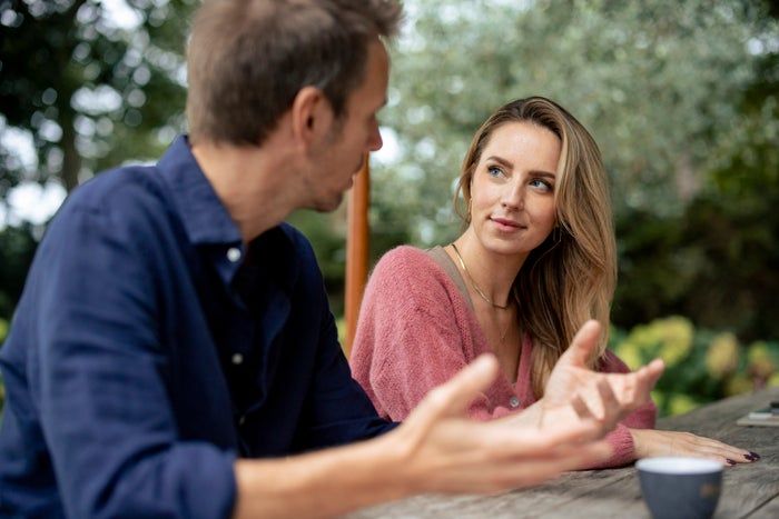 Two individuals seated at an outdoor table, conversing, one wearing a pink sweater, with a coffee cup in the foreground.