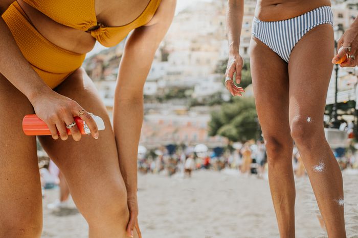 Two people at the beach applying sunscreen to their legs. One wears a striped bikini bottom, while the other wears a solid-colored bikini.