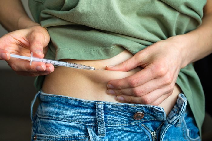 A person administering an injection into their abdomen, just above the waistband of their jeans.