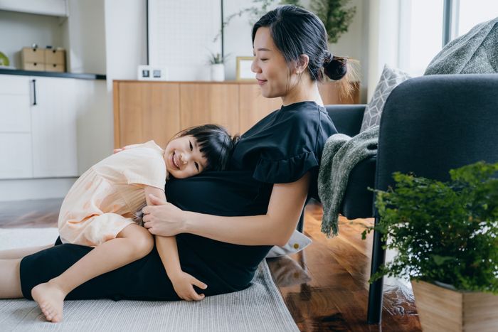 An image of a woman sitting on the floor with a child resting their head on the woman's pregnant belly, both smiling in a warm and inviting living room