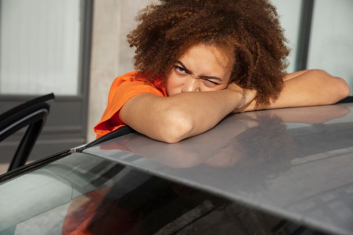 A person with curly hair resting their head on the roof of a car, squinting into the camera, wearing a casual top.