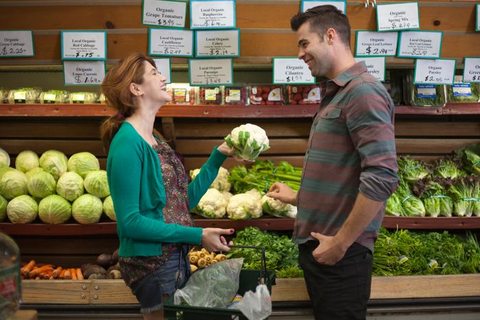 Two individuals in a grocery store; one is holding a cauliflower, both smiling beside a display of fresh vegetables.