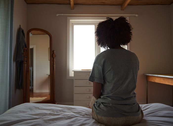 A person sits on a bed facing a window, with a mirror reflecting their image. The room feels serene and full of light.