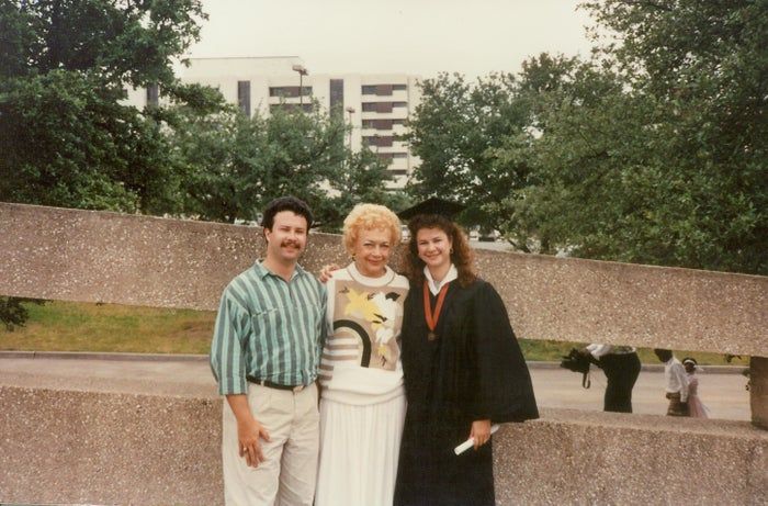 A man, an elderly woman, and a young woman in a graduation gown pose together outside, smiling for a photograph.