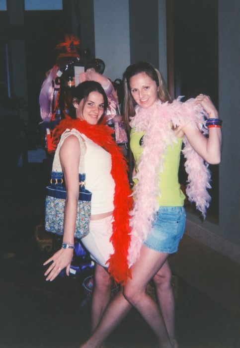 Two women are posing together, smiling widely and wearing feather boas — one in red and the other in pink. They seem to be thoroughly enjoying a lighthearted, casual event. Their names remain unknown.