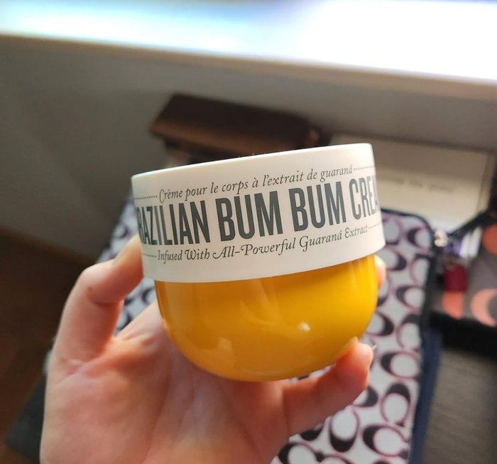 A hand holds a jar of Brazilian Bum Bum Cream, clearly showing its branding and text