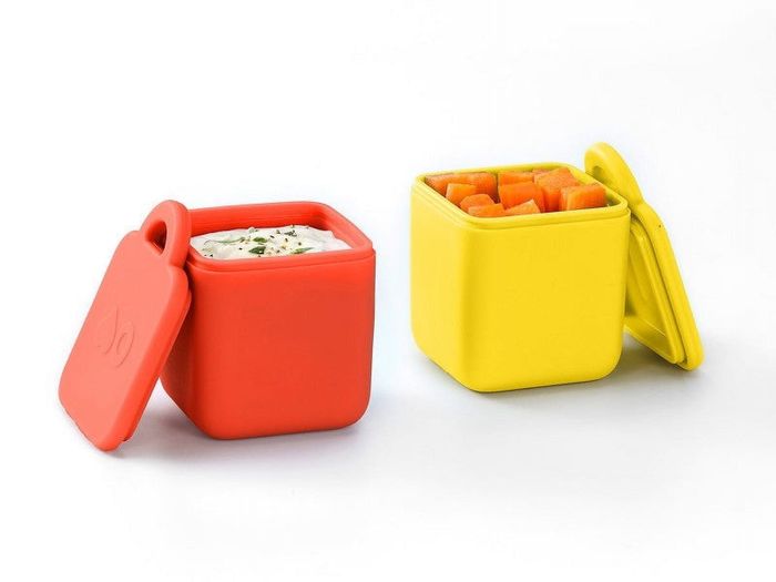 Two square silicone food containers, one red and one yellow, with lids partially open — one holding dip and the other filled with carrot sticks